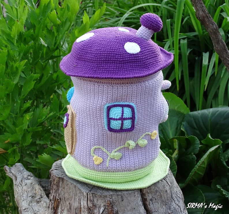 May include: A crocheted mushroom-shaped house with a purple roof, white polka dots, and a green base. The house has a small window with a blue frame and a light purple background. The house is sitting on a tree stump.
