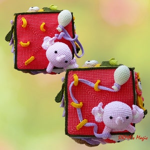 May include: Crocheted baby toy cube in red with yellow accents, featuring a lavender elephant and other colorful details. The cube has various textures and shapes, designed for sensory exploration and play. The toy is handmade and appears soft and safe for infants.