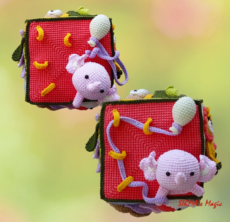 May include: Crocheted baby toy cube in red with yellow accents, featuring a lavender elephant and other colorful details. The cube has various textures and shapes, designed for sensory exploration and play. The toy is handmade and appears soft and safe for infants.