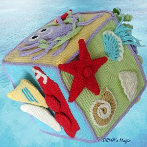 May include: A crocheted cube toy with different sea-themed designs on each side, including an octopus, a starfish, a seashell, and a surfboard. The cube is made in shades of blue, green, yellow, purple, and red.