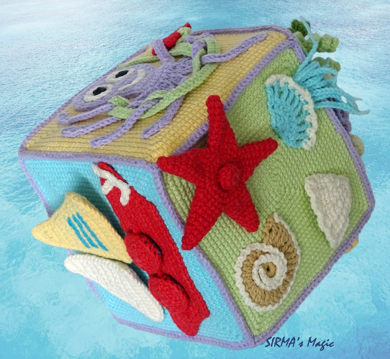 May include: A crocheted cube toy with different sea-themed designs on each side, including an octopus, a starfish, a seashell, and a surfboard. The cube is made in shades of blue, green, yellow, purple, and red.