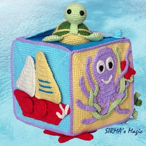May include: A crocheted cube toy with different ocean-themed designs on each side. The cube features a sailboat, an octopus, a turtle, and a starfish. The cube is made with colourful yarn and has a playful and whimsical design.