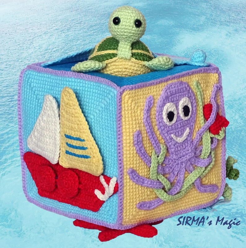 May include: A crocheted cube toy with different ocean-themed designs on each side. The cube features a sailboat, an octopus, a turtle, and a starfish. The cube is made with colourful yarn and has a playful and whimsical design.