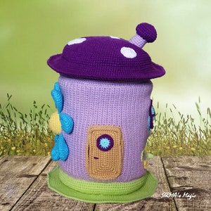 May include: A crocheted mushroom-shaped house with a purple roof, a lavender body, and a brown door with a blue peephole. The house has a green base and blue and yellow flower accents.