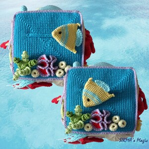 May include: Two crocheted squares with a blue background, featuring a yellow fish, coral, and seaweed. The squares are framed with a purple border and have a red crocheted border around the edges.