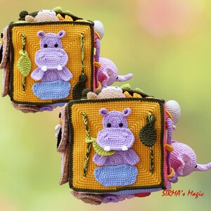 May include: Crocheted soft book featuring a purple hippo, blue water, and green leaf on a yellow square. The book has a brown border and includes other crocheted animals. The background is a soft green and yellow.