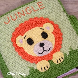 May include: A green crocheted baby book with the word "JUNGLE" on the cover. The page shown features a crocheted lion with orange mane, yellow face, and black eyes.