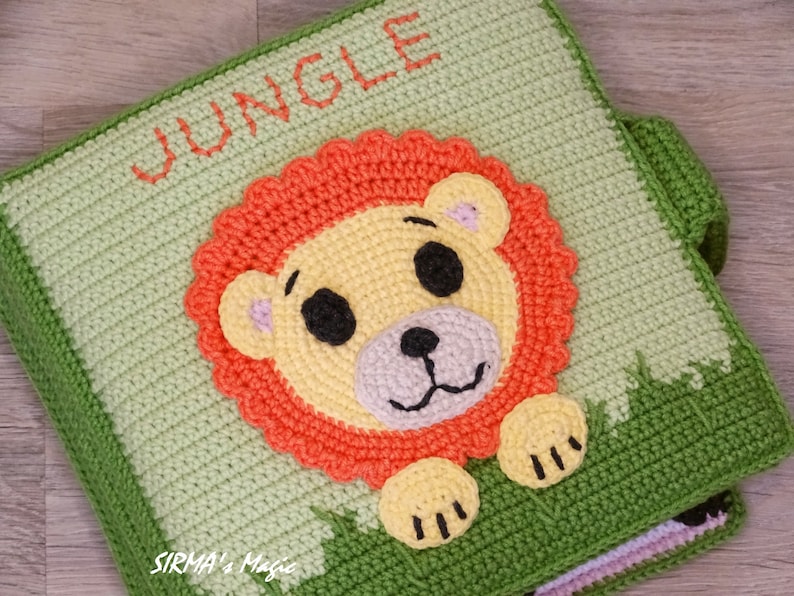 May include: A green crocheted baby book with the word "JUNGLE" on the cover. The page shown features a crocheted lion with orange mane, yellow face, and black eyes.