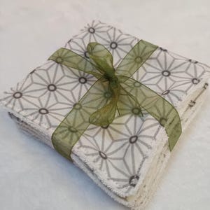 Puede incluir: Un conjunto de paños cuadrados de tela blanca con un patrón geométrico gris, atados con una cinta verde. Los paños están cuidadosamente apilados y parecen suaves, adecuados para diversos usos. La cinta añade un toque decorativo.