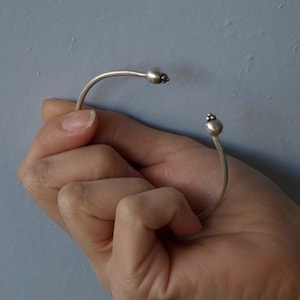 Könnte beinhalten: Ein silbernes Armreif-Armband mit zwei kleinen runden Perlen an den Enden.