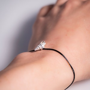 Puede incluir: Un brazalete de cordón negro con un dije de concha de mar plateado.