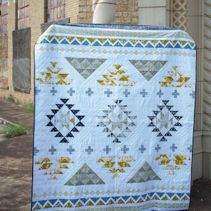 Puede incluir: Una colcha blanca con un patrón geométrico en tonos azules, amarillos y grises. La colcha presenta un diseño repetitivo de triángulos y cuadrados. La colcha está colgada en una pared de ladrillo.