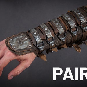 May include: A pair of brown leather and metal arm guards with buckles and rivets. The guards are worn on a person's arm and have a metal plate on the wrist.
