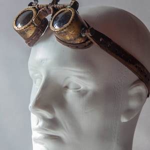 Op de afbeelding: Een paar steampunk-brillen met een bruin leren bandje. De bril heeft een messing frame en heldere glazen.