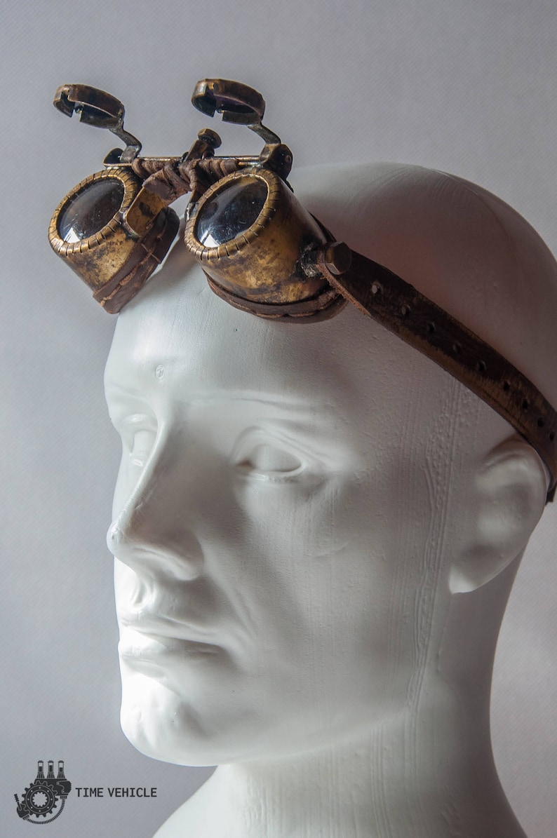 Op de afbeelding: Een paar steampunk-brillen met een bruin leren bandje. De bril heeft een messing frame en heldere glazen.