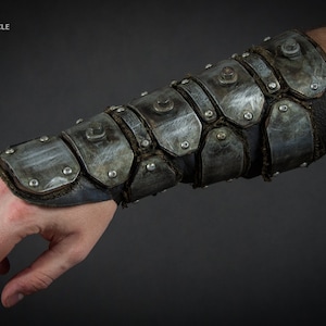 May include: A close-up of a metal arm guard with a distressed finish. The guard is made of multiple metal plates that are riveted together. The guard is worn on a person's forearm.