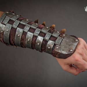 May include: A metal and leather forearm guard with a grid pattern and rivets. The guard is worn on a person's left arm.