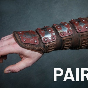 May include: A pair of brown leather arm guards with metal plates and rivets. The guards are worn on a person's forearm. The word "PAIR" is written in white letters on a dark grey background.