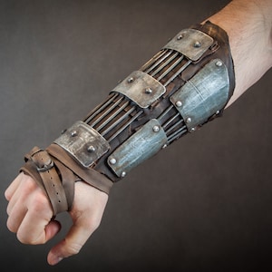 Könnte beinhalten: Ein Unterarm-Schutz aus Metall und Leder mit einem Riemenverschluss. Der Schutz besteht aus einer Reihe von Metallplatten und Riemen und ist dazu bestimmt, den Arm des Trägers zu schützen.