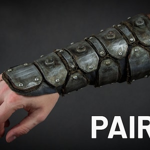 May include: A pair of metal arm guards with a distressed finish. The guards are made of multiple metal plates connected by rivets and straps. The guards are worn on the forearm and wrist.
