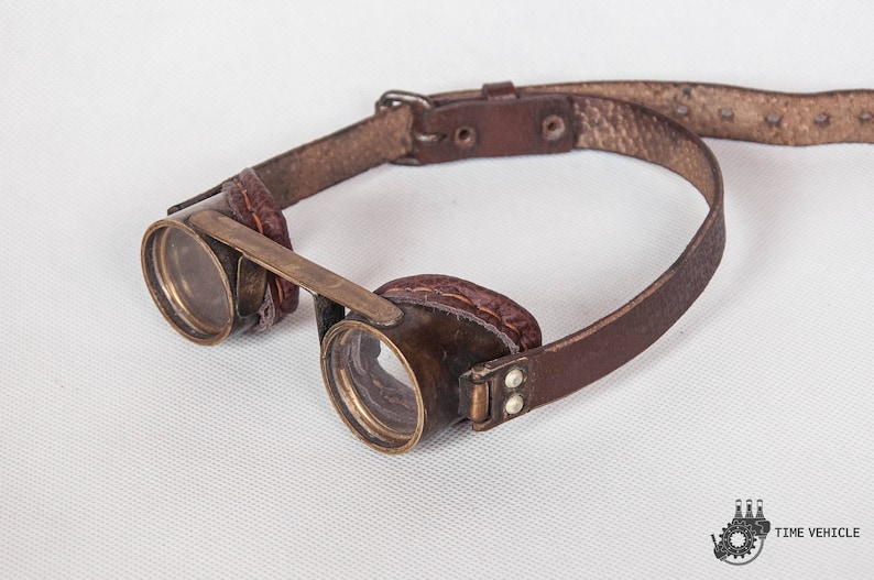 May include: A pair of steampunk-style goggles with round brass lenses and a brown leather strap. The goggles have a brass frame and are held together by a brass bar.