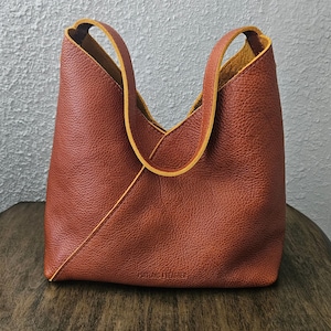 May include: A rust-colored leather hobo bag with a pebbled texture, featuring a diagonal seam across the front, two integrated shoulder straps, and the embossed text PORTLAND LEATHER at the bottom center of the exterior surface.