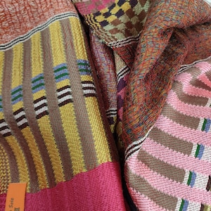 May include: A colourful woven fabric scarf with a pink, yellow, brown and blue geometric pattern. The scarf has a tag that reads "100% Soie".
