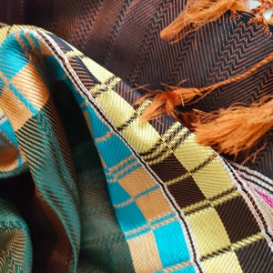 May include: A brown, black and turquoise plaid scarf with orange tassels. The scarf is made of a soft, woven fabric.