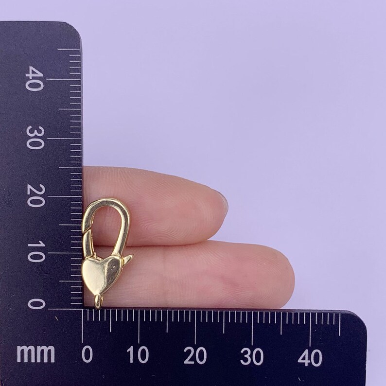 May include: Gold-toned heart-shaped lobster clasp for jewellery making. The clasp is approximately 1.5 cm long and is shown next to a ruler.