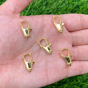 May include: Five gold-coloured metal lobster clasps, also known as trigger snaps, are shown on a hand. These clasps are commonly used for attaching straps, chains, or other accessories.