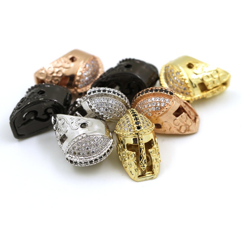 May include: A collection of gold, silver, black, and rose gold gladiator helmet charms with clear and black rhinestones. The charms are designed to be used for jewelry making.
