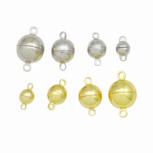 May include: A collection of eight spherical magnetic clasps arranged in two rows. The top row features four silver-toned clasps in descending sizes, while the bottom row displays four gold-toned clasps in ascending sizes, each with two loops.