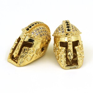 May include: Two gold-tone gladiator helmet charms with black and clear rhinestones. The charms are detailed with intricate designs and have a shiny finish.