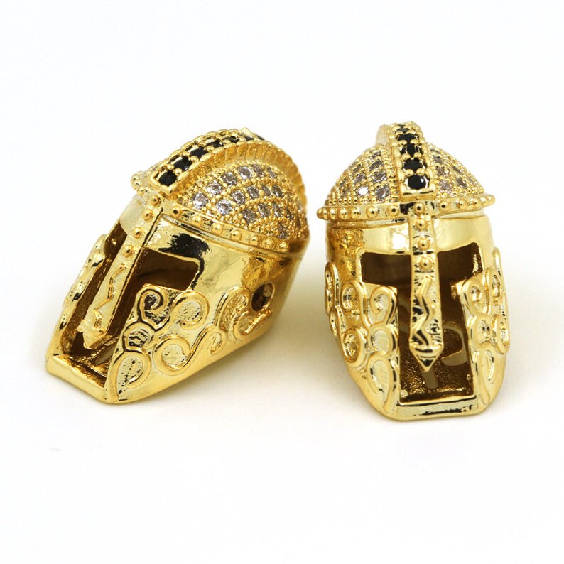 May include: Two gold-tone gladiator helmet charms with black and clear rhinestones. The charms are detailed with intricate designs and have a shiny finish.