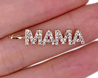 Puede incluir: Colgante dorado con la palabra "MAMA" en mayúsculas. Cada letra está incrustada con pequeños cristales transparentes, creando un efecto brillante. El colgante tiene un pequeño lazo para sujetarlo a una cadena.