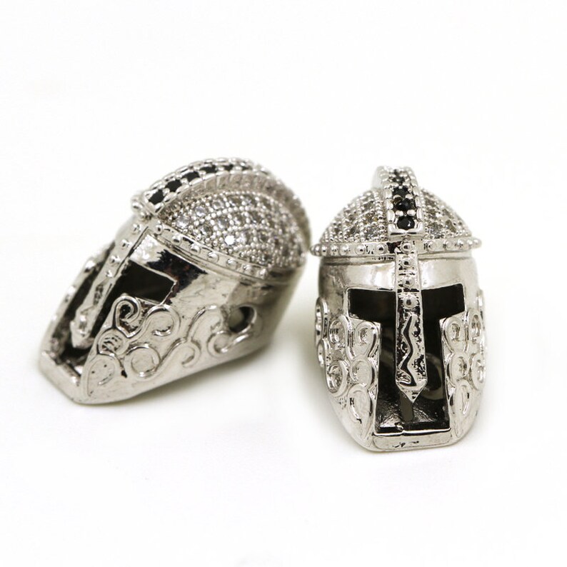 May include: Two silver-toned helmet charms with intricate details and black and white rhinestones.
