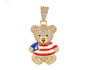 Puede incluir: Colgante de oso de peluche de tono dorado con un diseño de bandera americana de esmalte rojo, blanco y azul. El oso está cubierto de piedras de circonita cúbica transparentes.