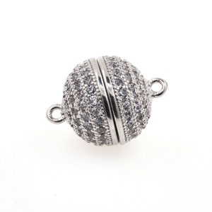 Op de afbeelding: Een bolvormige magnetische sluiting voor sieraden met een zilverkleurige afwerking, voorzien van een pav&eacute;-zetting van talloze kleine, heldere, rondgeslepen stenen die het oppervlak bedekken. De sluiting heeft twee kleine ringetjes aan de zijkanten.