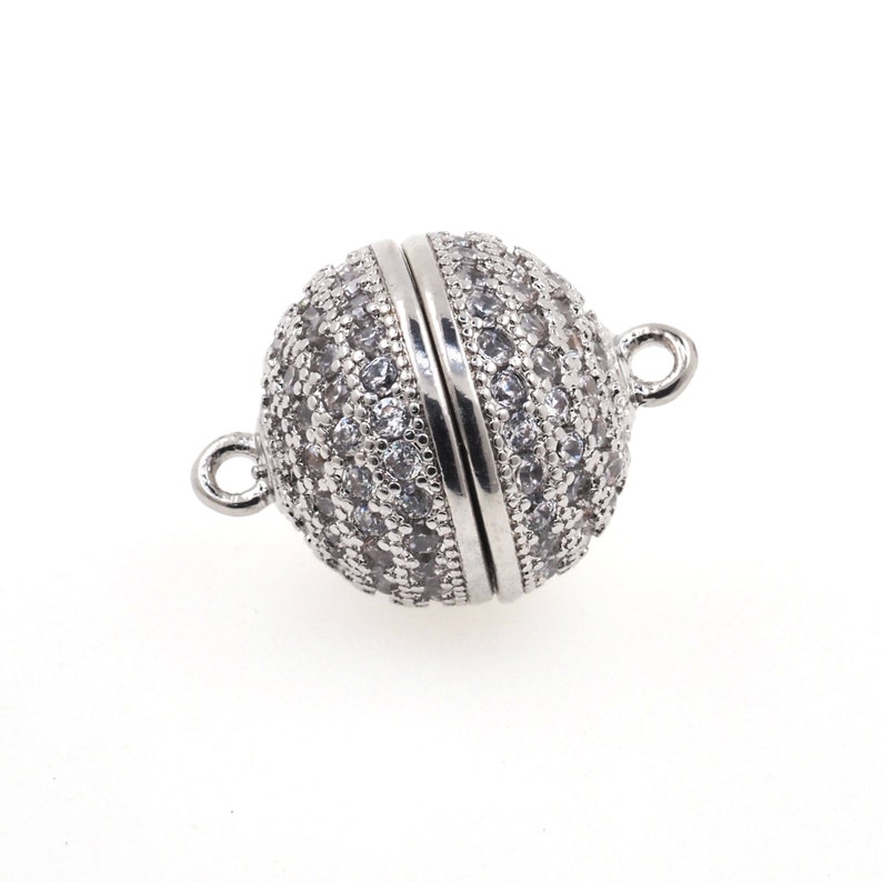 Op de afbeelding: Een bolvormige magnetische sluiting voor sieraden met een zilverkleurige afwerking, voorzien van een pav&eacute;-zetting van talloze kleine, heldere, rondgeslepen stenen die het oppervlak bedekken. De sluiting heeft twee kleine ringetjes aan de zijkanten.