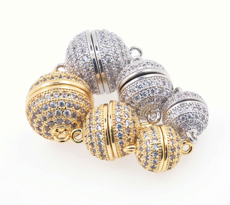 Op de afbeelding: Een collectie bolvormige magnetische sluitingen voor sieraden in goud- en zilverkleuren. Elke ronde sluiting is ingelegd met talloze kleine, heldere, sprankelende steentjes en heeft een klein metalen oogje aan de boven- en onderkant.