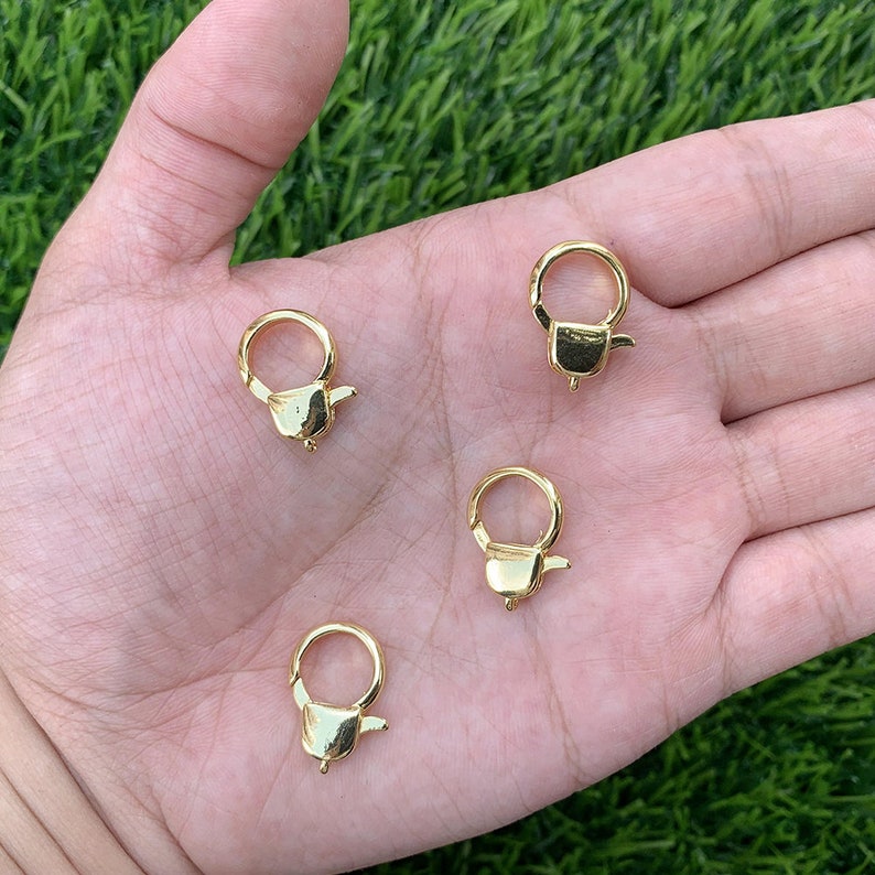 May include: Four gold-toned metal lobster clasps, also known as swivel clasps, are shown on a hand. These clasps are commonly used for jewellery making and crafting.