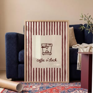 Puede incluir: Impresión artística enmarcada con una ilustración de una cafetera y el texto "coffee o'clock". La obra de arte tiene un fondo crema con rayas verticales burdeos. El marco es de madera clara. La impresión se muestra en un salón.
