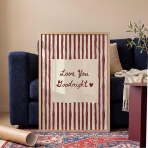 Op de afbeelding: Een ingelijste print met een licht houten frame. De print heeft een crèmekleurig vierkant met de woorden "Love You Goodnight" in een scriptlettertype, met een hartsymbool. De achtergrond heeft verticale bordeauxrode strepen.