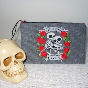 May include: Gray zippered pouch with an embroidered design of two skeletons holding each other, surrounded by red roses and a banner that reads "Endless Love".