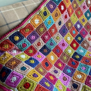 Pode incluir: Uma manta de crochê colorida e feita à mão com um design de retalhos de quadrados em várias cores vibrantes. A manta tem uma borda rosa e é drapeada sobre um sofá bege. Os quadrados são preenchidos com padrões florais.