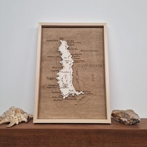 May include: A wooden map of Lundy Island, a small island off the coast of Devon, England. The map is framed in a light wood frame and features a detailed outline of the island with various landmarks and geographical features labeled in cursive script. The map is mounted on a light brown wood background.