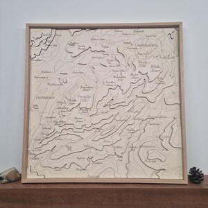 May include: A laser-cut wooden map of the Pendle Hill area in Lancashire, England. The map features the names of various towns, hills, and moors, including Pendle Hill, Clitheroe, and Nelson. The map is framed in a light wood frame.
