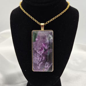 May include: A gold-toned necklace featuring a rectangular pendant with a purple and green marbled design. The pendant is encased in a gold frame and hangs from a delicate gold chain. The necklace is displayed on a black velvet bust.