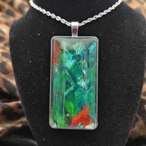 May include: Silver-toned necklace with a rectangular pendant featuring an abstract design. The pendant showcases a vibrant mix of green, red, and white colors, encased in a clear resin. The chain is a delicate, linked design.