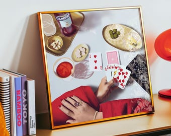 May include: A framed art print with a gold frame. The artwork depicts a table setting with playing cards, drinks, and snacks. A hand holds playing cards with hearts. The color palette includes red, gold, and white.